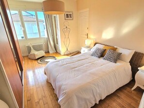 Un dormitorio con una cama grande, una mesita de noche con lámpara, un radiador y una ventana con cortinas.