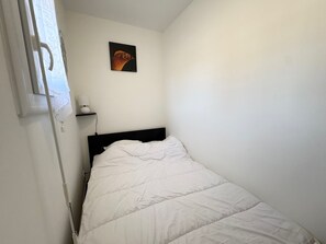 Ein kleines Zimmer mit einem Einzelbett, einer Bettdecke in Weiß, einem Nachttisch mit Lampe und einem an der Wand hängenden Bild.