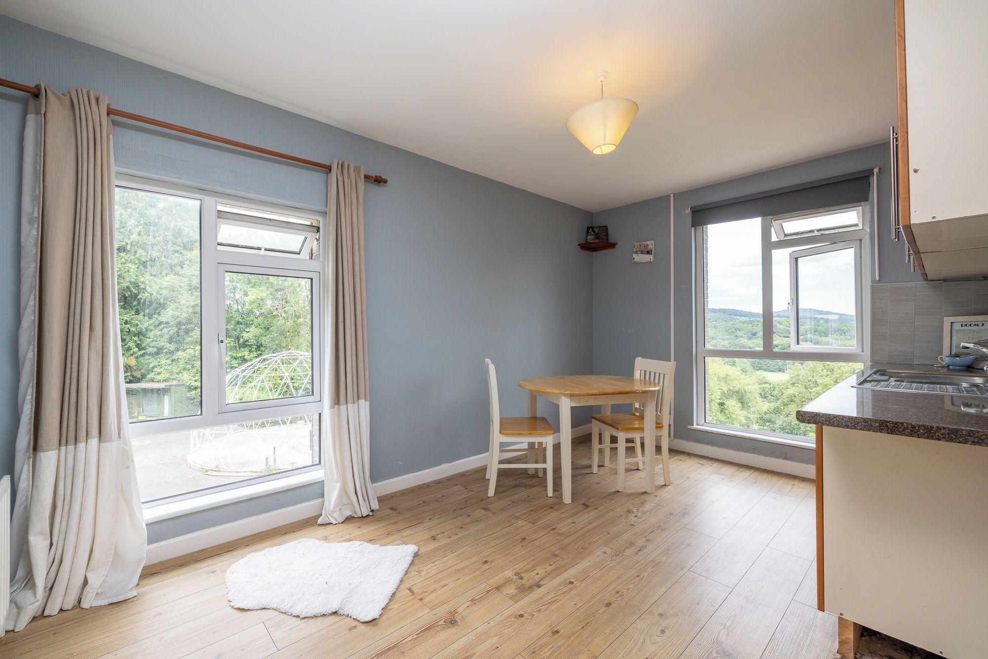 Een kamer met een eettafel en stoelen, grote ramen met gordijnen, een wit vloerkleed en een keukenhoek.