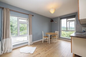 Quarto com mesa de jantar e cadeiras, janelas amplas com cortinas, tapete branco e área de cozinha.