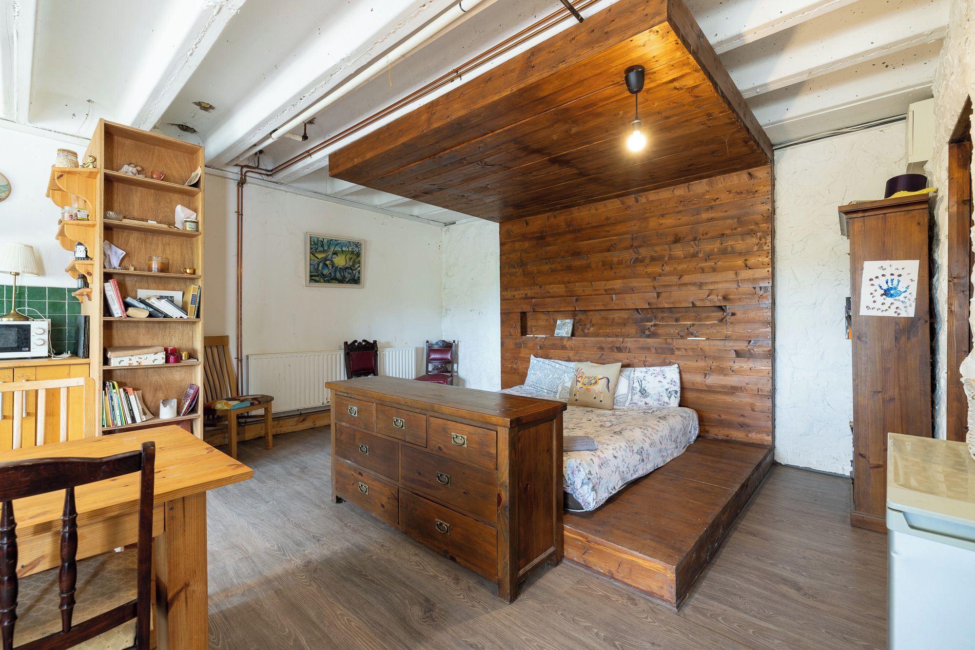 A room with a wooden ceiling, a bed with patterned bedding, a wooden dresser, and a bookshelf.
