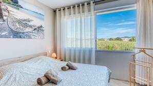 A bedroom with a bed, a window with a view, a painting on the wall, and a wooden drying rack. - Apartment with AC, sea view - 4 guests (Le Lavandou)