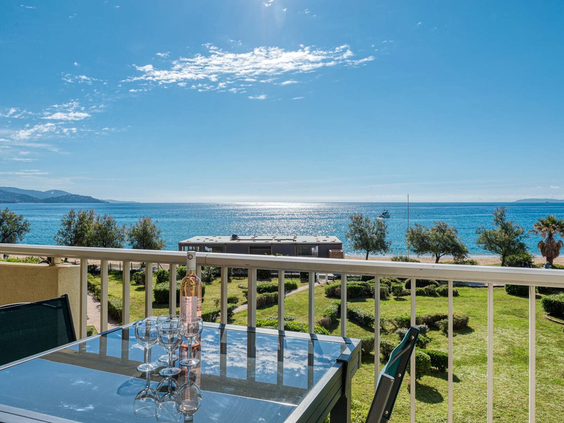 A balcony with a glass table, wine glasses, and a bottle, overlooking a clear blue sea and a distant shoreline.