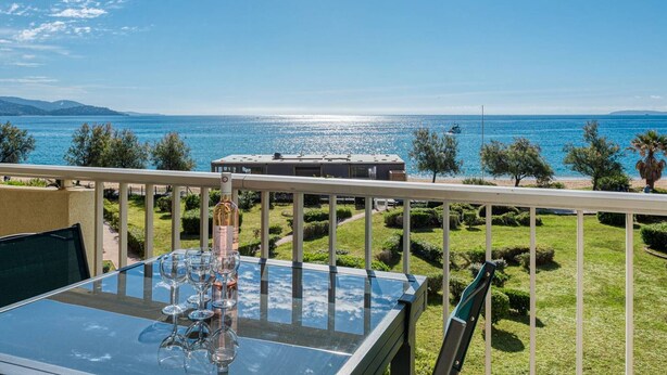 A balcony with a glass table, wine glasses, and a bottle, overlooking a clear blue sea and a distant shoreline. - Apartment with AC, sea view - 4 guests (Le Lavandou)