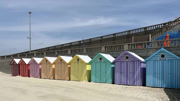 Eine Reihe bunter Strandhäuser mit Sandstrand und einer Betonwand im Hintergrund.