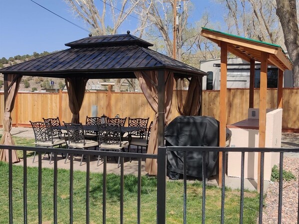 A covered patio with a table and chairs, a grill, and a wooden fence. - Cedar City RV Resort (Cedar City)
