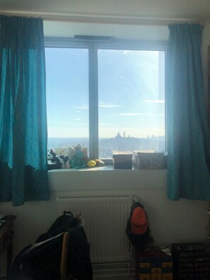 Ein Zimmer mit Fenster und Blick auf die Stadt, einem Heizkörper und einem Rucksack auf dem Boden.