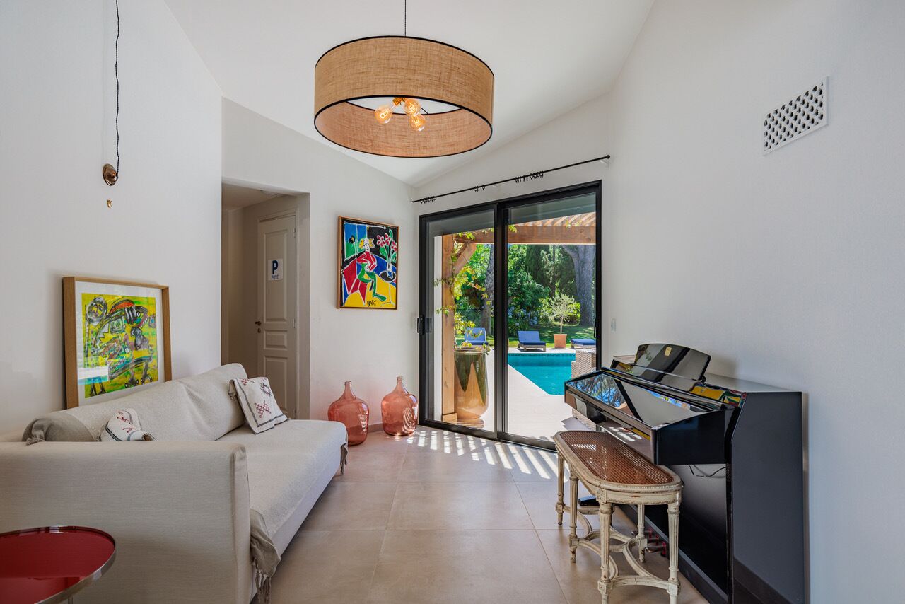 Una sala de estar moderna con un sofá, un piano, una mesa roja y un gran ventanal con vistas a una piscina.