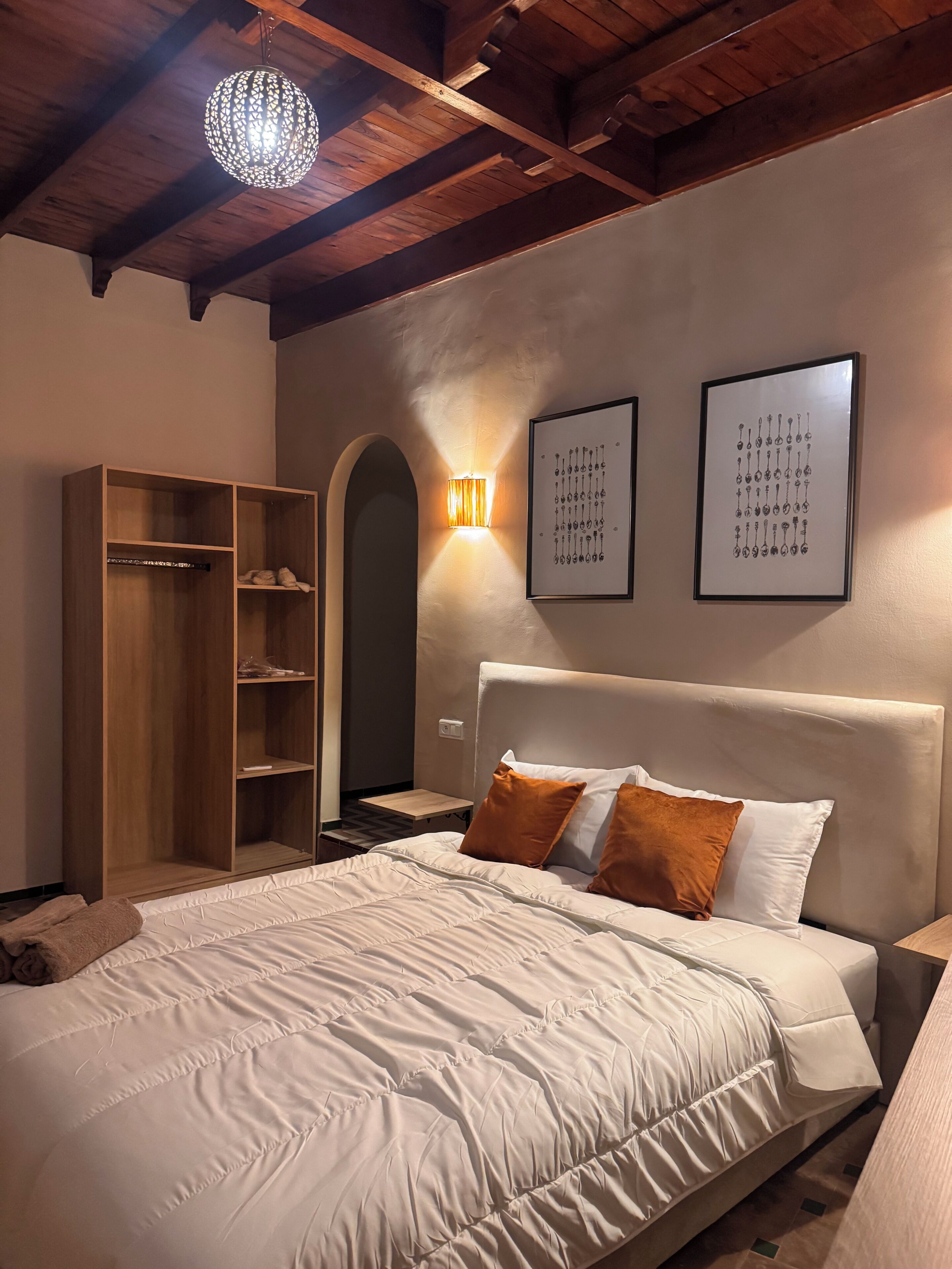 A bedroom with a bed, wooden wardrobe, wall-mounted light, and two framed pictures.