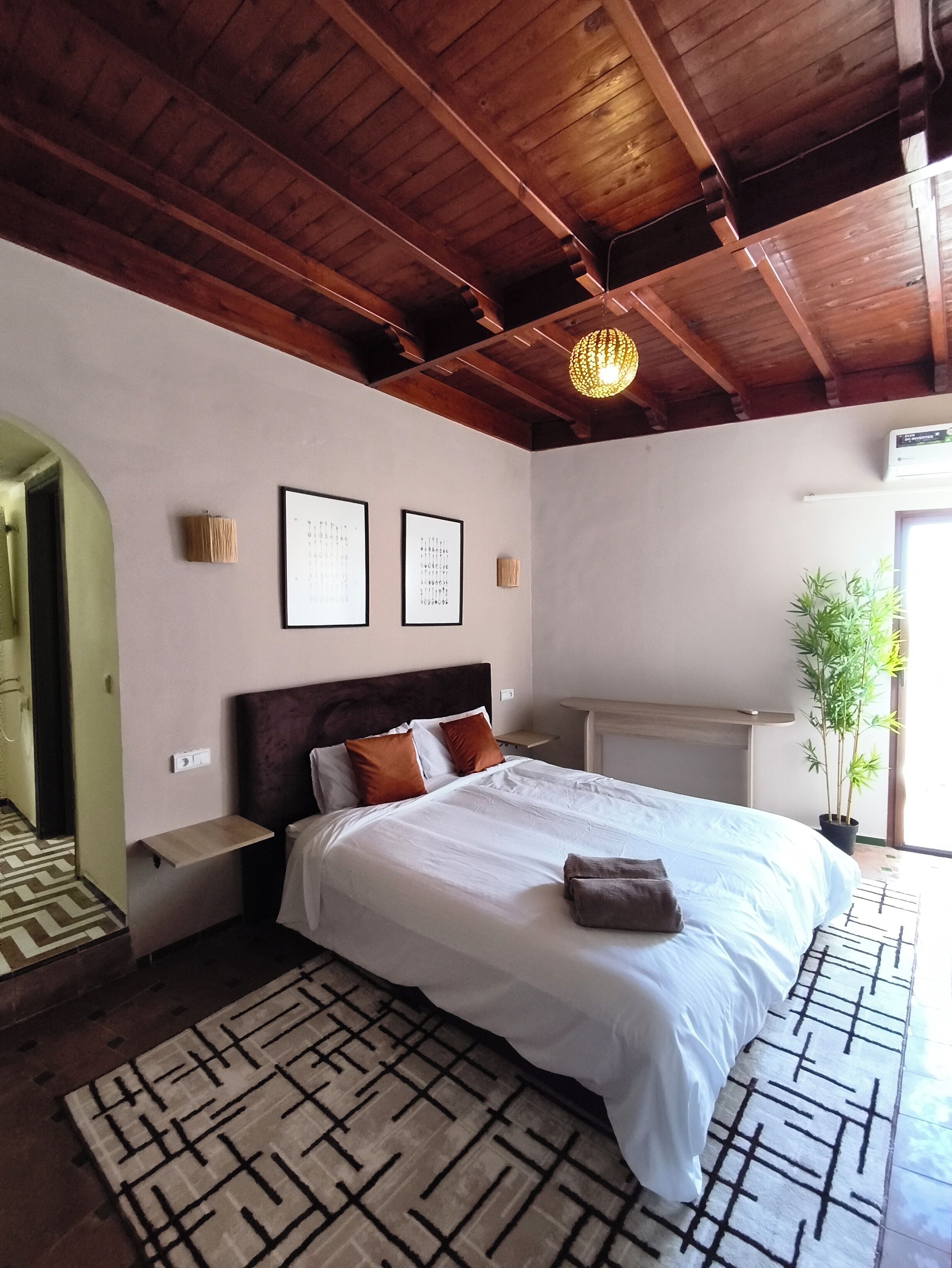 Un dormitorio con techo de madera, una cama con ropa de cama blanca, un tapete estampado y una planta en maceta.