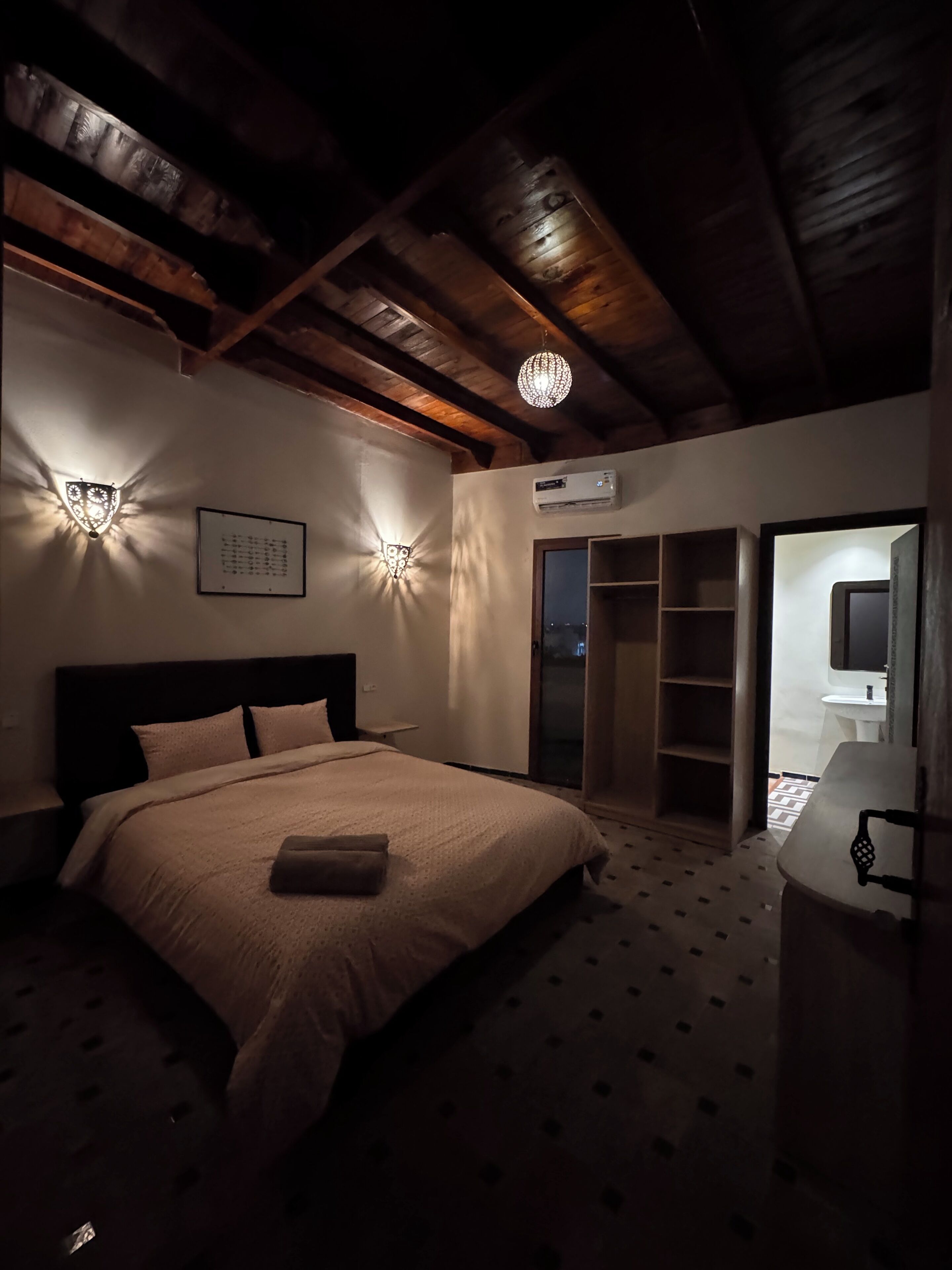 Un dormitorio con cama, apliques de pared, unidad de aire acondicionado, armario y baño con lavamanos y espejo.