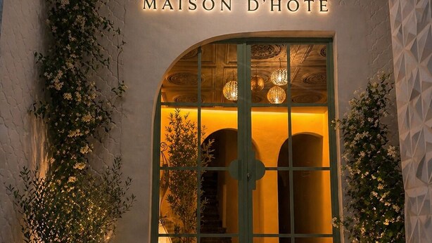 Entrada de hotel con un cartel que dice 'MAISON D'HÔTE', una puerta de cristal y plantas en macetas a ambos lados.