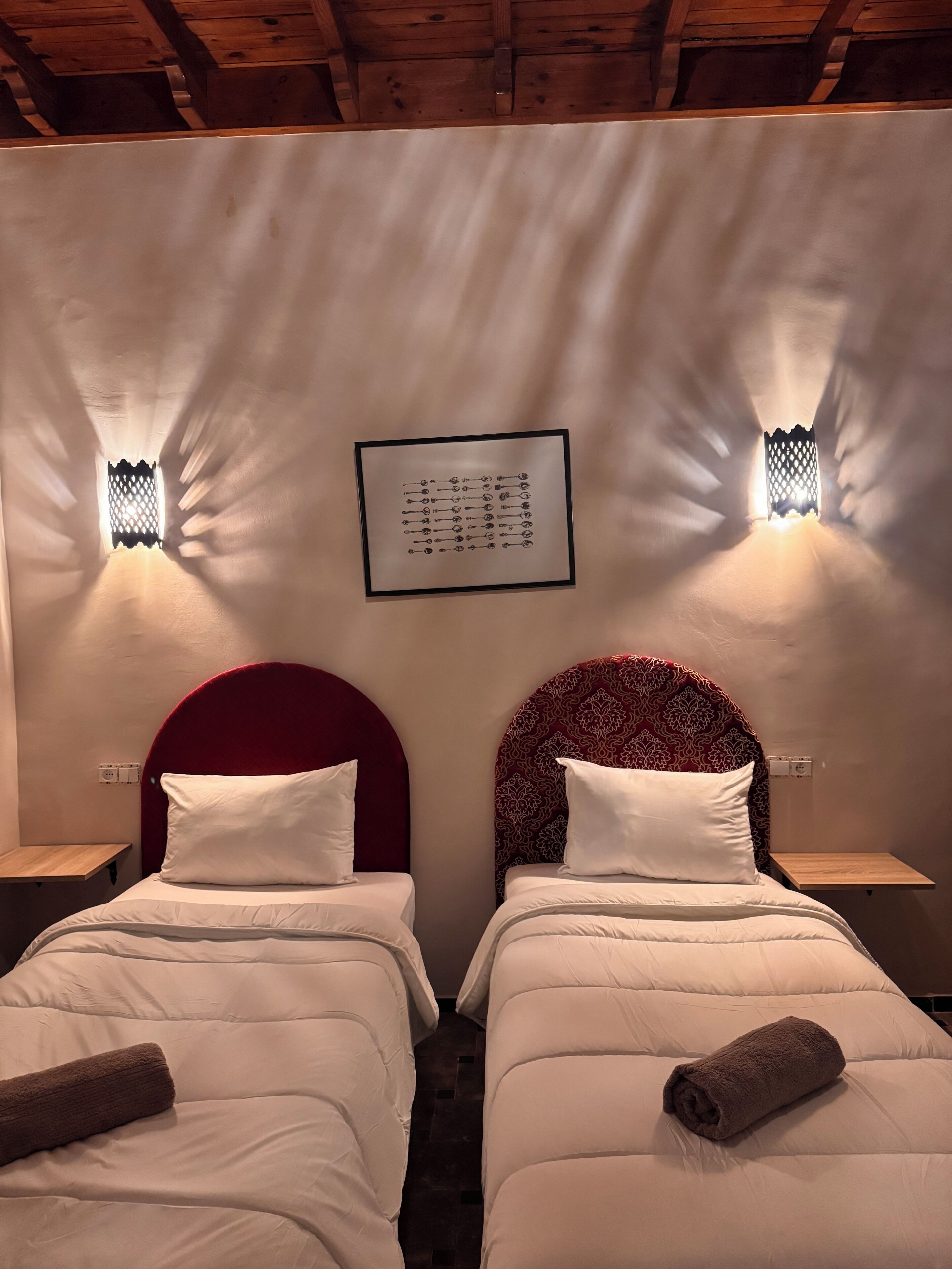A hotel room with two beds, each with a red headboard, white bedding, and a rolled towel. There are wall-mounted lights and a framed picture above the beds.