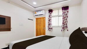 A bedroom with a bed, an air conditioning unit, a wooden door, and a window with floral curtains.