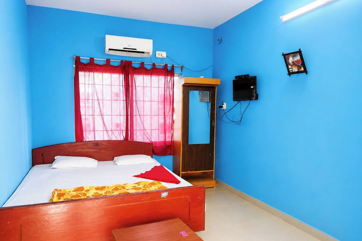 A room with a bed, a small table, a wall-mounted air conditioner, a door, and a wall-mounted TV.