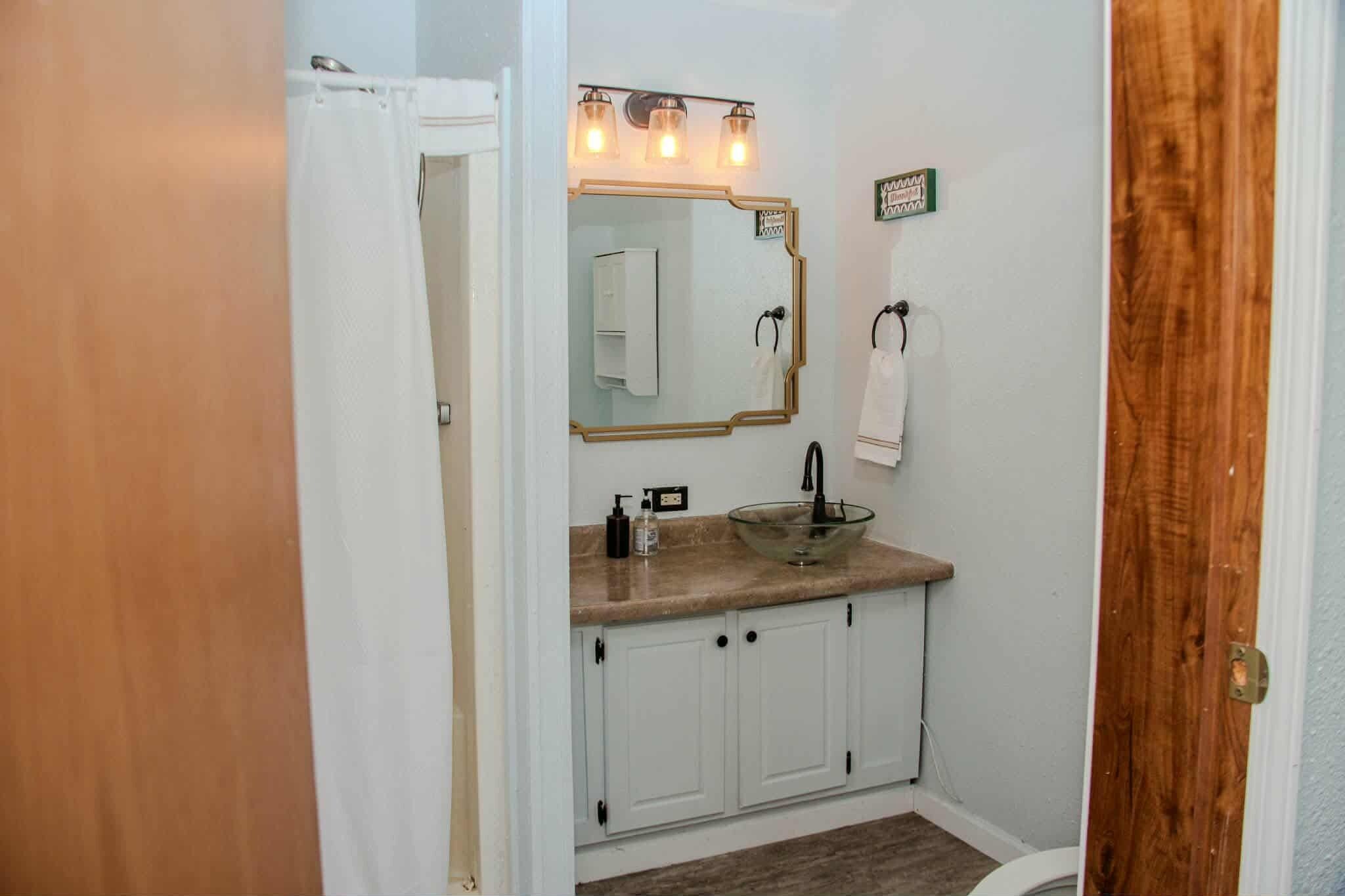 A bathroom with a white sink, a mirror with lights, a towel rack, and a wooden door.