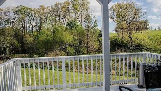 A covered outdoor area with a white railing, a grill, and a view of a grassy landscape with trees. - Easy Access Ramp Entry  3-bedroom 2 bathroom Harrison Cottage with AC (Harrison)