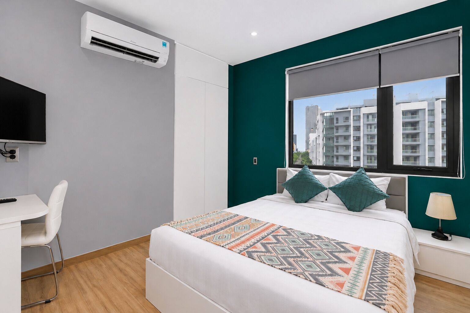 A modern hotel room with a bed, a desk, a chair, an air conditioner, a window with a view of buildings, and a lamp.