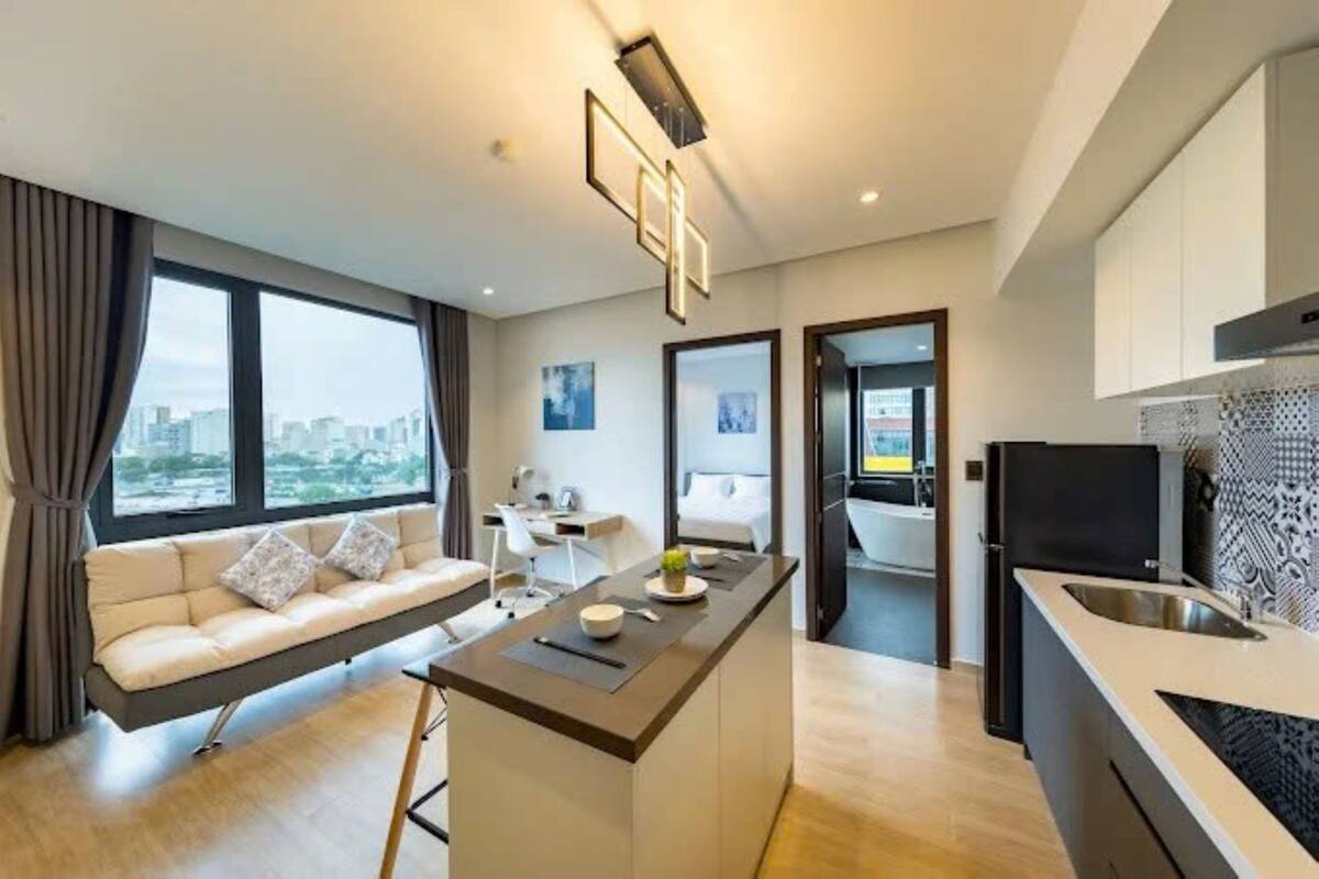 A modern kitchen with a central island, a dining area with a sofa and a TV, and a view of the city through large windows.