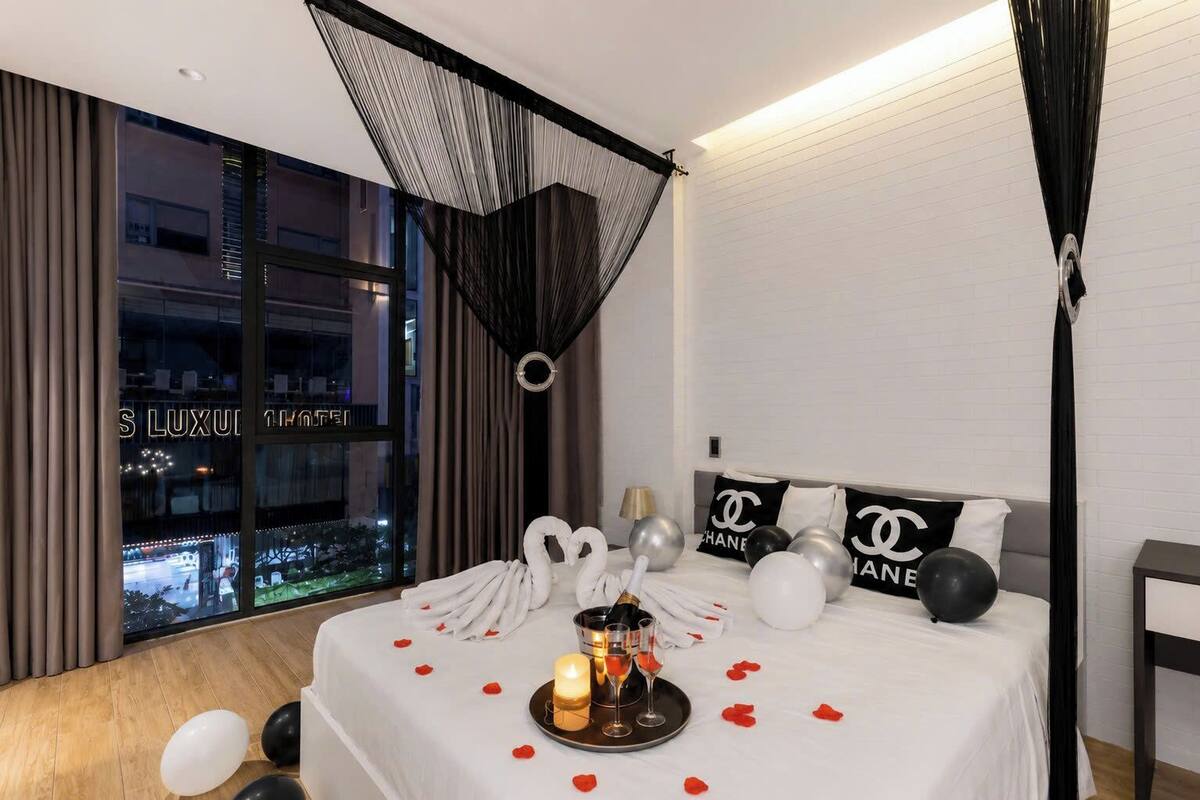 A hotel room with a bed, a chandelier, a view of the city, and Chanel-themed pillows.