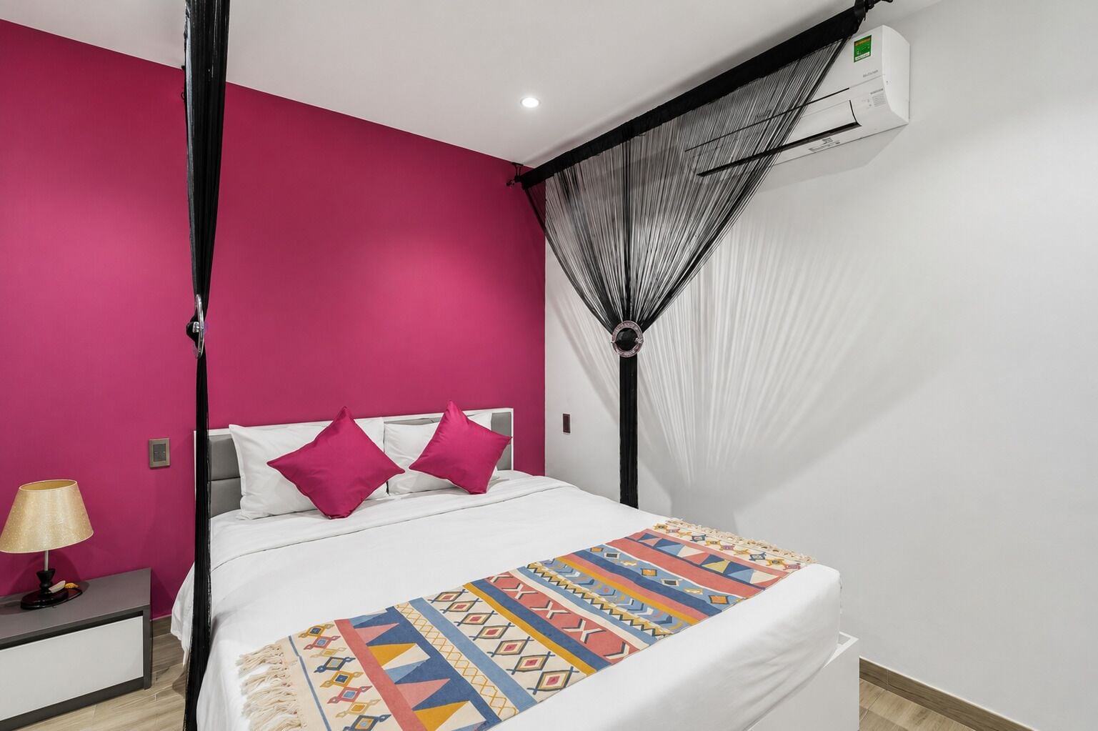 Una habitación de hotel con cama, mesita de noche, lámpara, mosquitero y aire acondicionado.