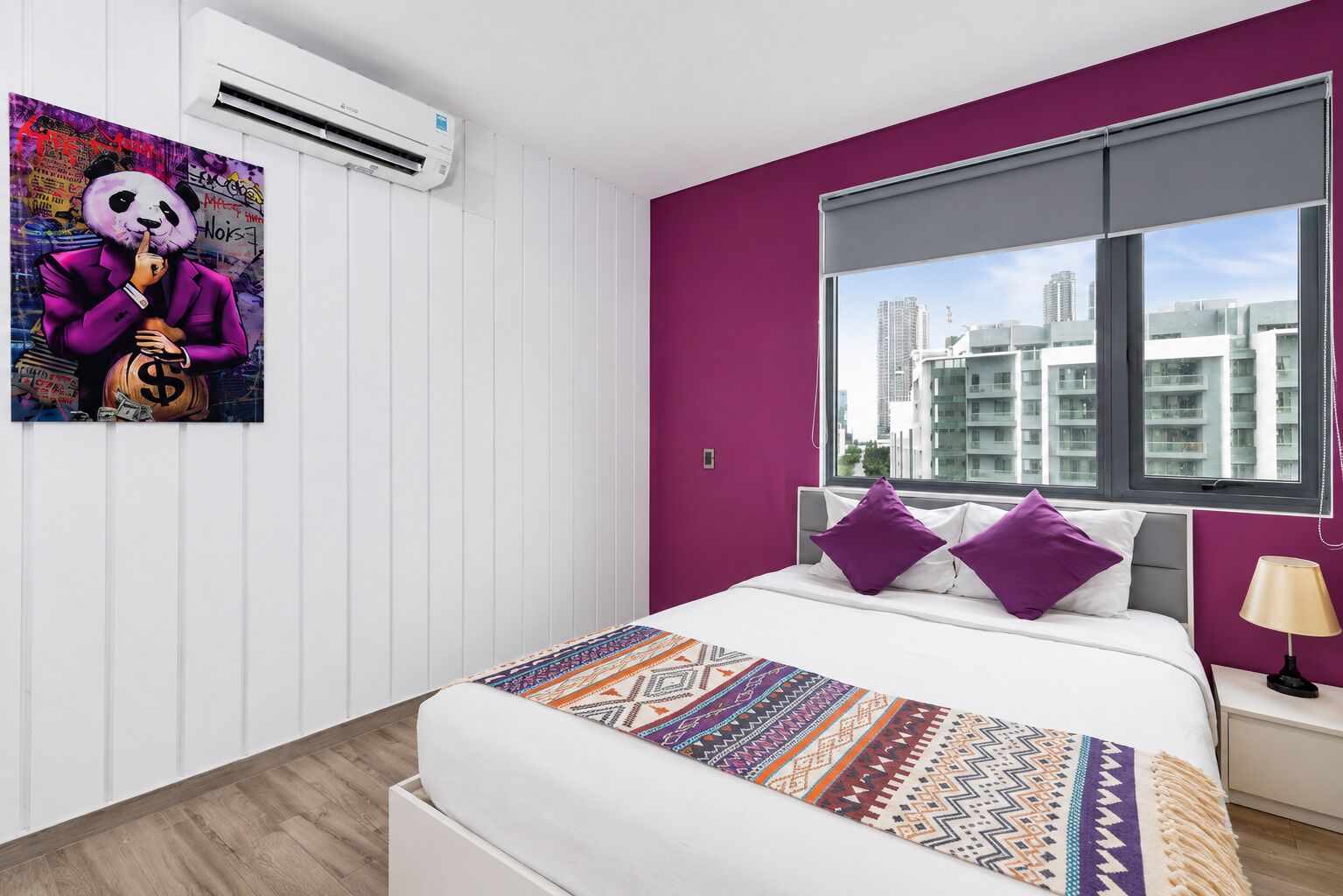 A modern bedroom with a city view, a bed with patterned bedding, and a wall-mounted air conditioner.