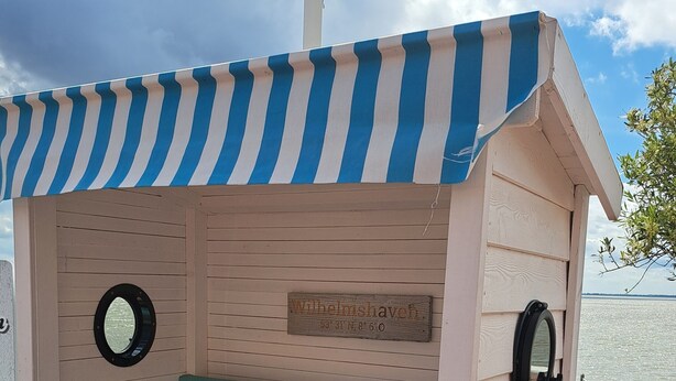 Une maison de plage avec un auvent rayé, une plaque nominative et une vue sur l’eau.