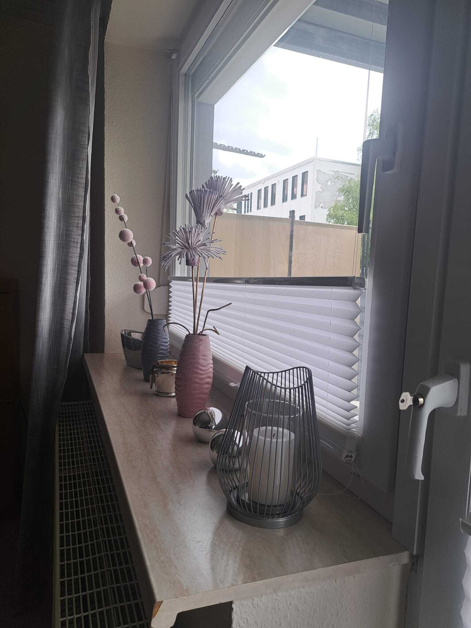 Ein Balkon mit Fenster, einem Metallstuhl und Dekorationselementen wie Vasen und einer Pflanze.