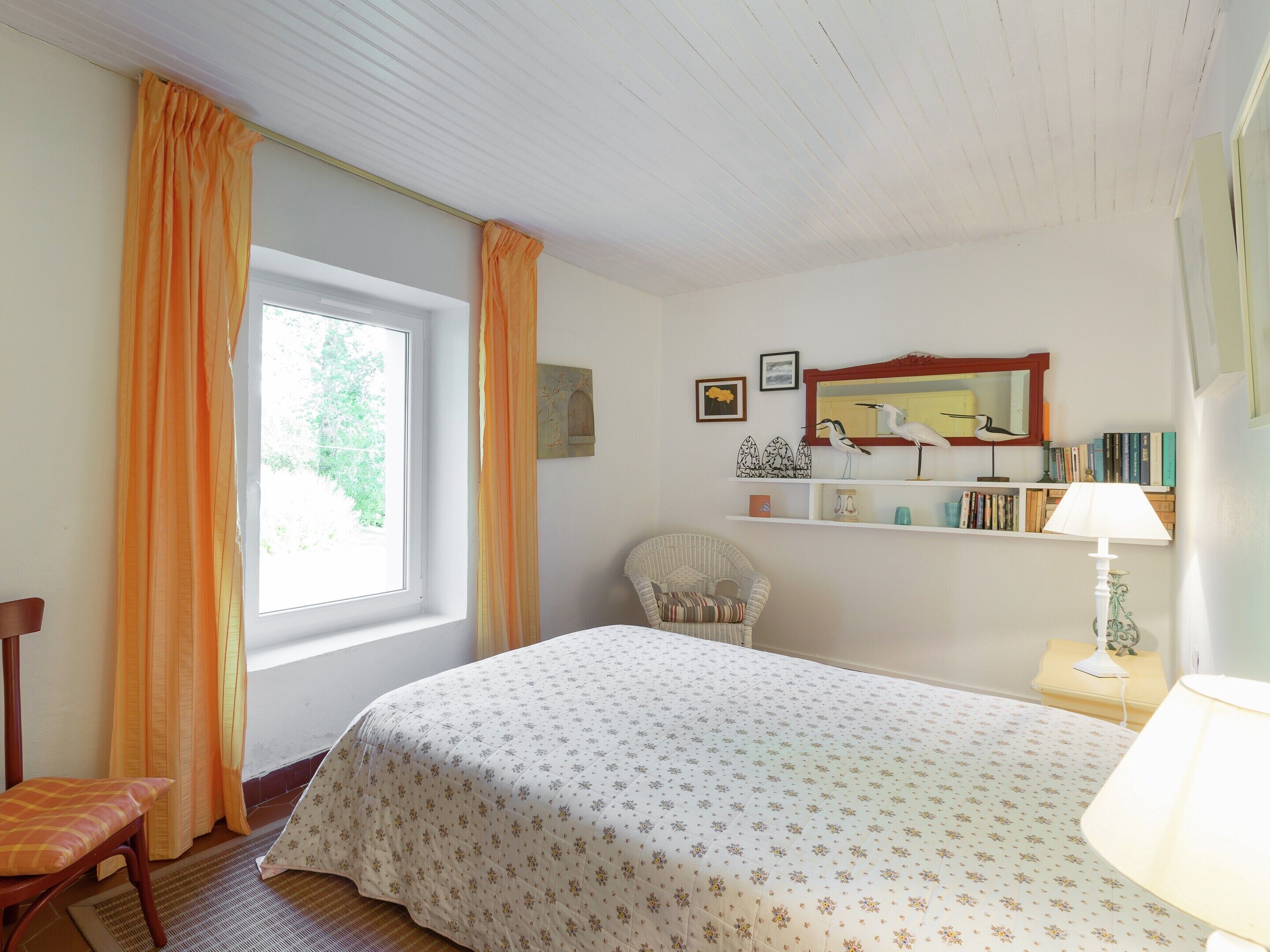 Un dormitorio con una cama, una silla, una ventana con cortinas, una estantería con libros y una lámpara.