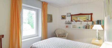 Un dormitorio con una cama, una silla, una ventana con cortinas, una estantería con libros y una lámpara.