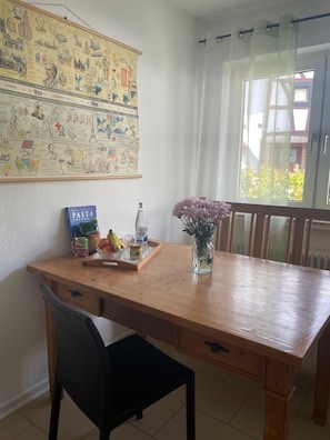 Uma área de refeições com uma mesa de madeira, uma cadeira, um livro de receitas de massas, uma garrafa e um vaso com flores.