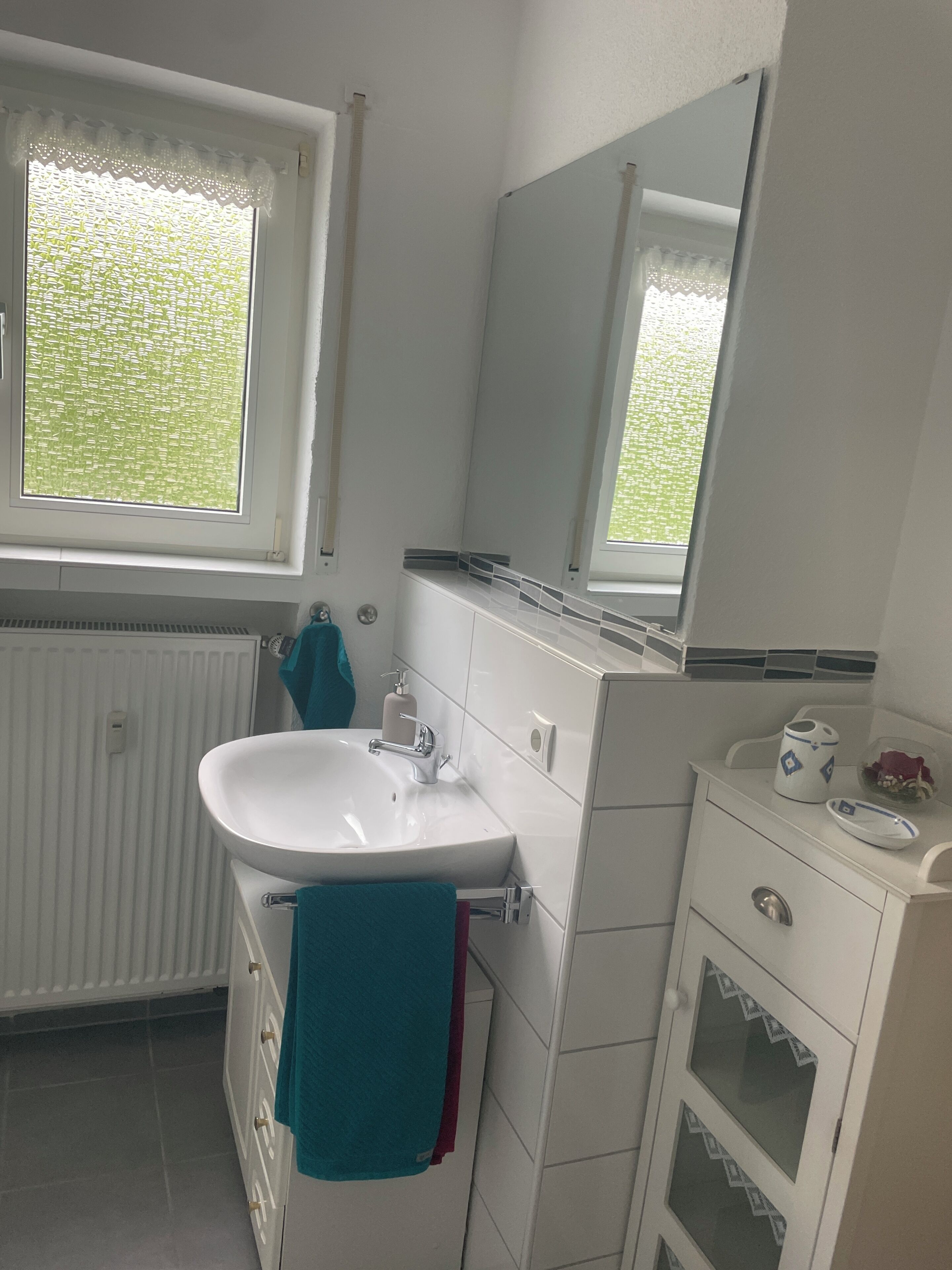 Een badkamer met een witte wastafel, een spiegel, een raam met uitzicht op groen en een witte radiator.