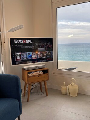 Un soggiorno con televisione che mostra l'interfaccia di Netflix, un tavolino in legno con libri, una poltrona blu e vista mare attraverso un'ampia finestra.