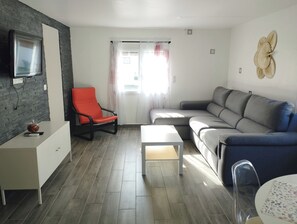 Un salon moderne avec un canapé gris, un fauteuil rouge, une table blanche et un téléviseur fixé à un mur en briques.