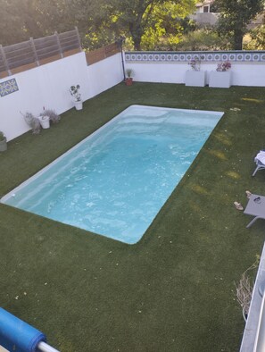 Piscina retangular rodeada por relva sintética, com um muro branco e azulejos decorativos ao fundo.