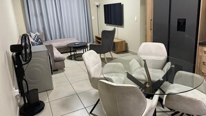 A modern living room with a sofa, a glass table with chairs, a TV, a refrigerator, and a fan. - Rivonia Modern 2Bed Apartment (Sandton)
