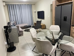 A modern living room with a sofa, a glass table with chairs, a TV, a refrigerator, and a fan.