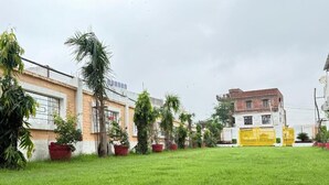 A grassy area with buildings in the background, including a yellow gate and a building with a sign. - Hotel JB Square Ayodhya Near Ram Mandir, Airport & Ayodhya Cantt (Faizabad)