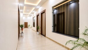 A hallway with a wooden floor, potted plants, and a large window with dark curtains. - Hotel JB Square Ayodhya Near Ram Mandir, Airport & Ayodhya Cantt (Faizabad)