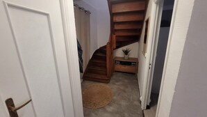 Un pasillo con una escalera de madera, una puerta, una alfombra pequeña y una planta sobre un estante de madera.