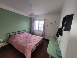 Une chambre à coucher comprenant un lit, une table de chevet, une fenêtre avec des rideaux, un radiateur et une étagère fixée au mur.