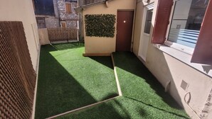 Un patio estrecho con césped artificial verde, un banco de madera, una puerta y una pared cubierta de plantas trepadoras.