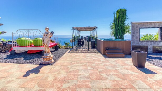 En patio med statue, seng med grønne puder, pergola og havudsigt.