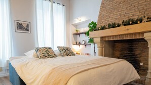 A bedroom with a bed, a fireplace, a window with curtains, and a wall-mounted shelf with plants. - Normandy Cottage City Center charming apartment (Évreux)