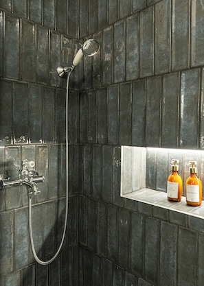 Une douche équipée d’un pommeau de douche à main et d’un robinet mural. Deux bouteilles de shampoing sont posées sur une étagère.