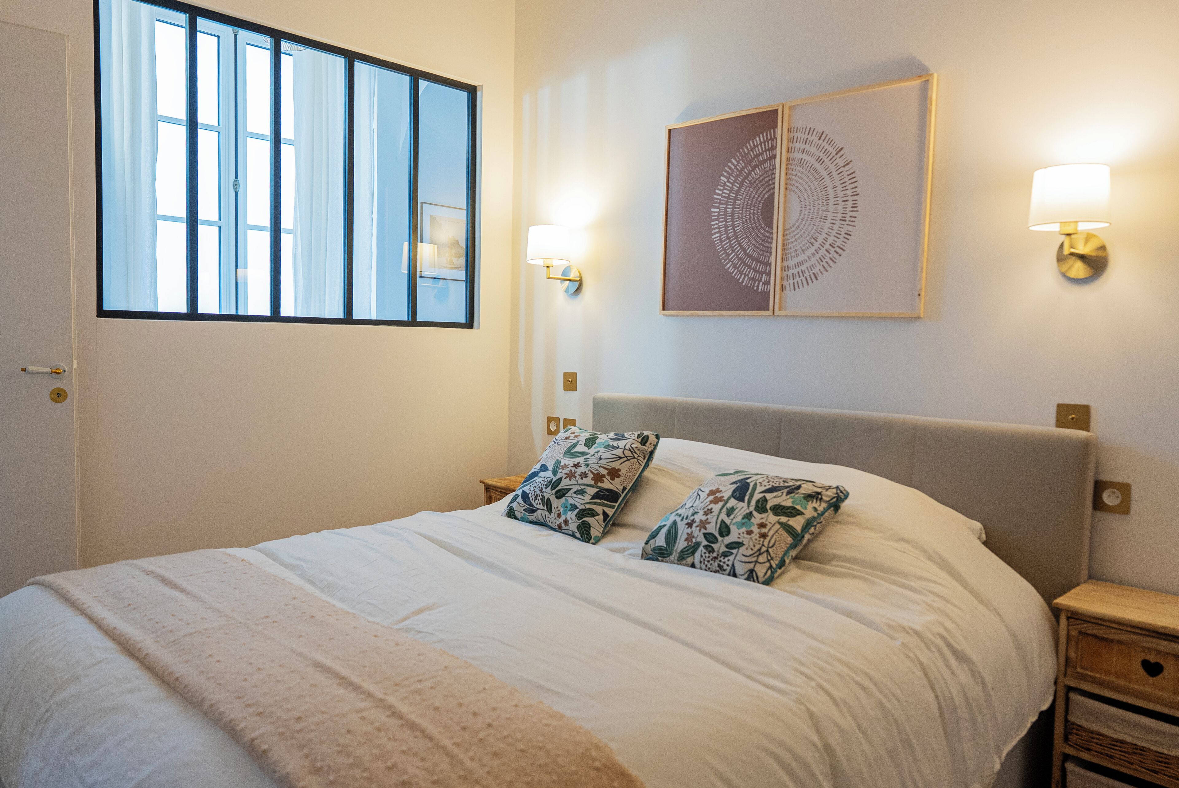 A neatly made bed with a white comforter and two decorative pillows, a bedside table with a lamp, and a wall-mounted artwork.