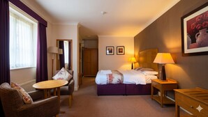 Une chambre d’hôtel comprenant un lit, deux tables de chevet avec des lampes, une chaise, une table ronde, un miroir et une porte donnant sur une autre pièce.