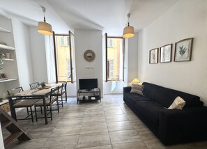 Una sala de estar con un sofá negro, una mesa de comedor con sillas, un mueble para la televisión y dos ventanas con contraventanas abiertas.