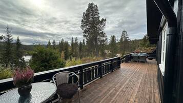 A wooden deck with a glass table and chairs, overlooking a forested area and a body of water.