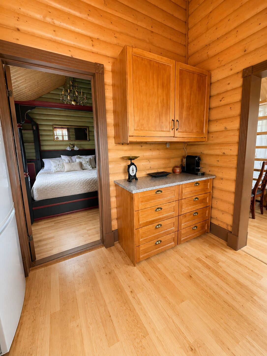 A wooden cabin interior with a bed, a kitchen area with cabinets and a scale, and a dining area with a chair.